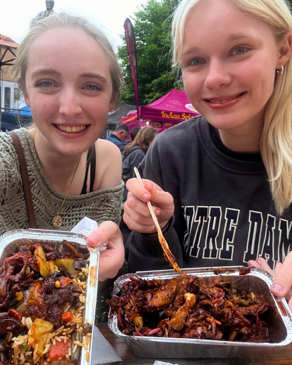 Two young women holding trays filled with food at an outdoor food market with colorful tents in the background.