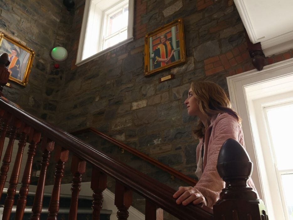Person ascending a wooden staircase inside a stone-walled building with framed artwork and a window letting in natural light.