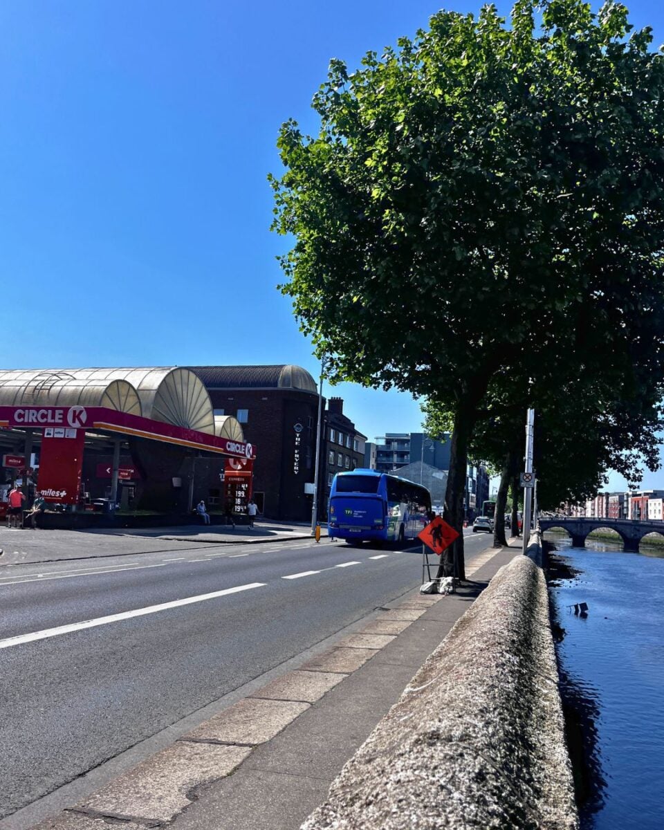 A sunny urban street scene with a Circle K gas station on the left and a large tree shading the sidewalk beside a river on the right.