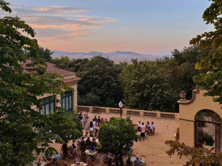 Outdoor terrace with people dining and socializing under a colorful sunset sky surrounded by trees and historic buildings.