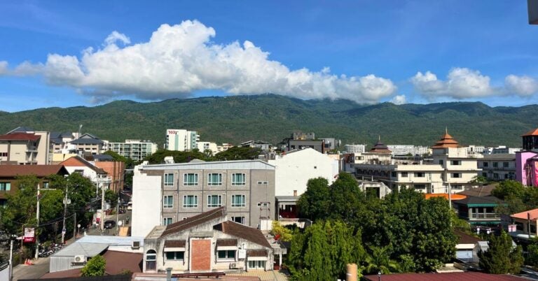 A cityscape with buildings and a backdrop of green mountains under a blue sky with clouds.