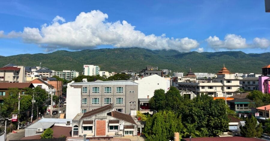 A cityscape with buildings and a backdrop of green mountains under a blue sky with clouds.