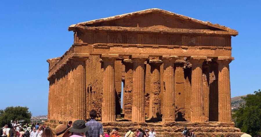 Ancient Greek temple with weathered columns under a clear blue sky, surrounded by tourists. The scene conveys a sense of historical exploration.