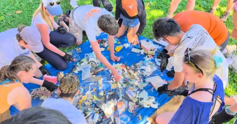 Group of people gathered outdoors on grass, sorting and collecting various pieces of trash on a blue tarp during a cleanup event.