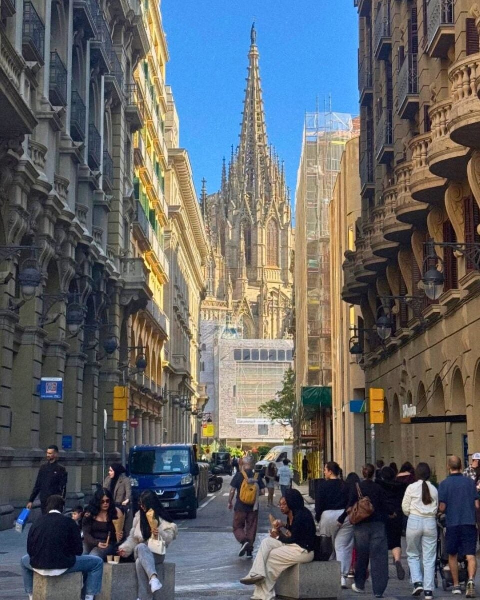 Narrow city street bustling with pedestrians and vehicles, framed by ornate historic buildings leading to a towering Gothic cathedral spire under a clear blue sky.