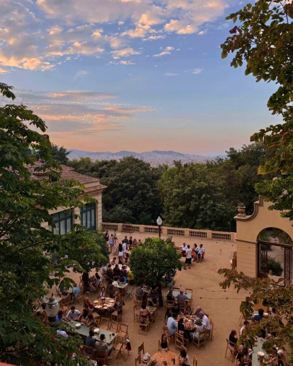 Outdoor terrace with people dining and socializing under a colorful sunset sky surrounded by trees and historic buildings.