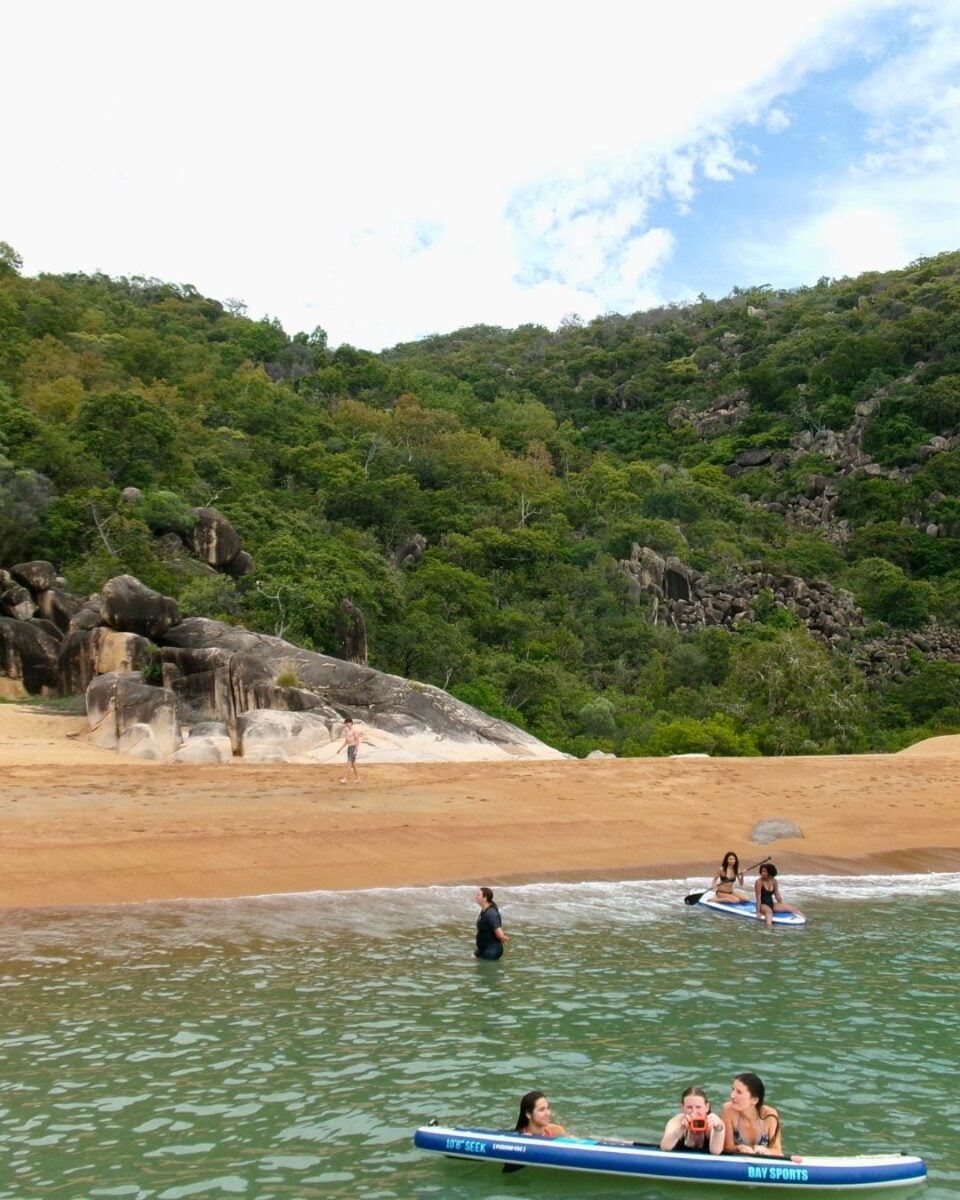 Beach scene with people on paddleboards and a lush, green hillside in the background.