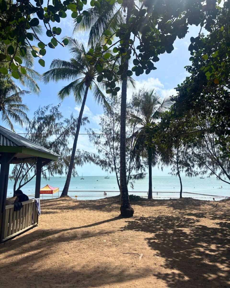Beachfront scene with palm trees framing the view of a calm sea, small boats, and a lifeguard tent on the sand.
