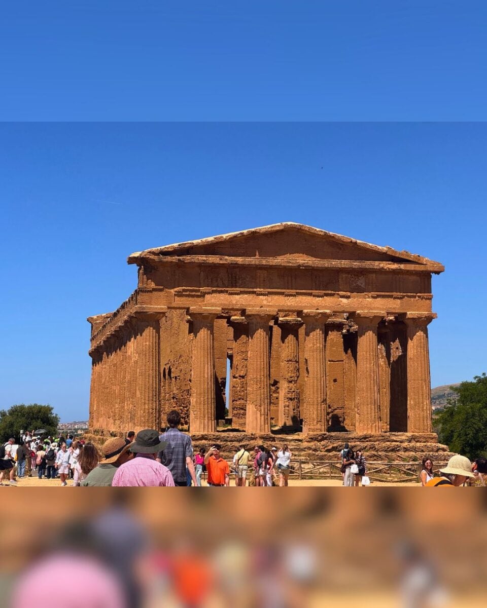 Ancient Greek temple with weathered columns under a clear blue sky, surrounded by tourists. The scene conveys a sense of historical exploration.