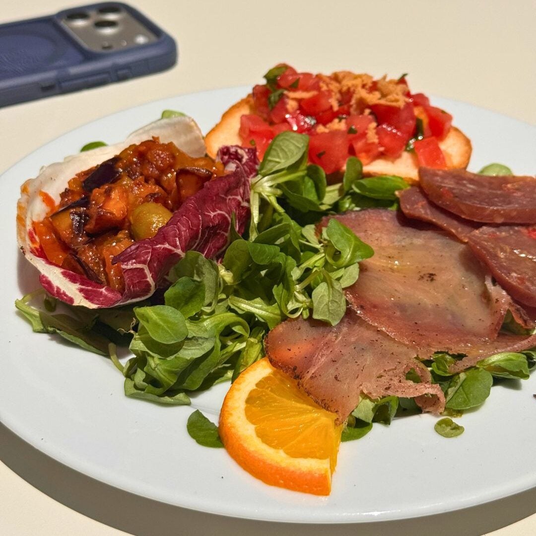A white plate with radicchio, vegetable mixture, cured meats, greens, bruschetta, and an orange slice. A smartphone is in the background.
