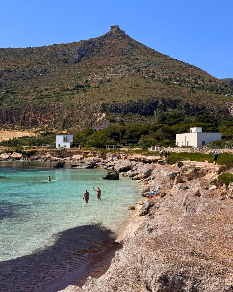 Coastal bay with turquoise water, rocky shore, and a hill with a stone ruin and white buildings.
