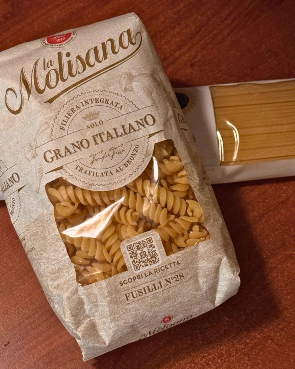 two packages of pasta on a wooden surface. In the foreground is a beige paper bag of fusilli pasta with a clear plastic window displaying the spiraled pasta inside. To the right, partially visible and placed horizontally, is a second package made of clear plastic containing spaghetti.