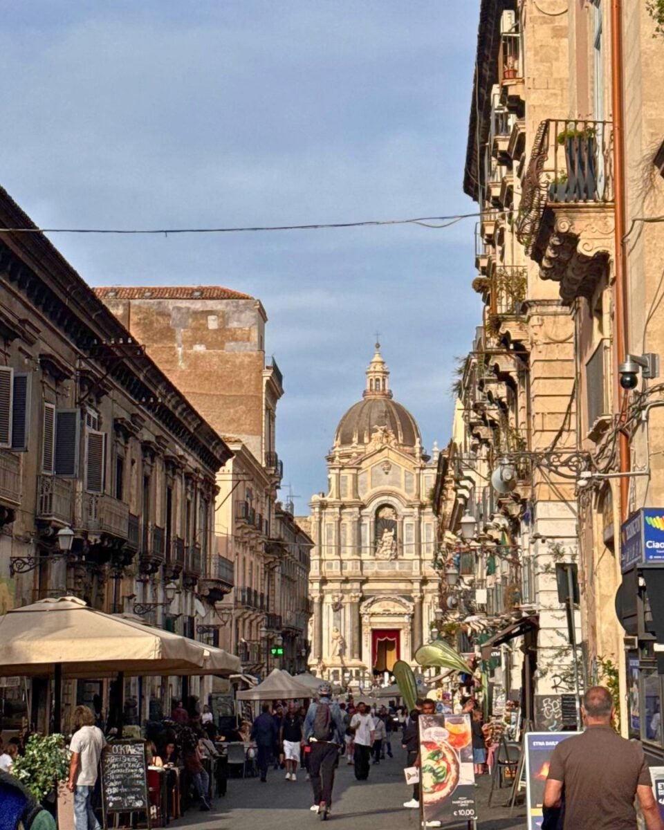 A bustling street in a historic city with cafes, shops, and a stunning baroque church in the background under a clear sky.