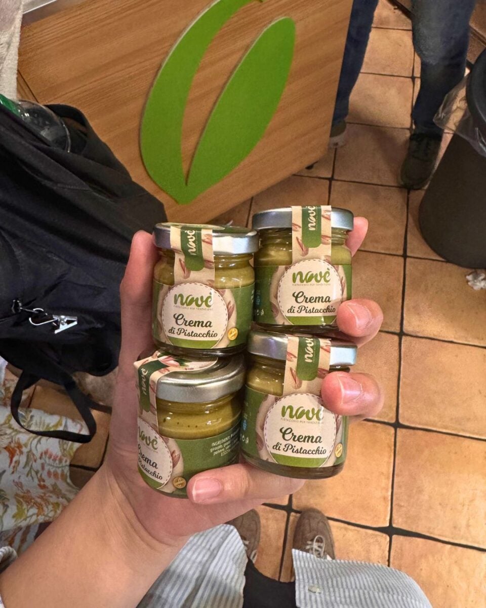 A hand holding four jars of Novi Crema di Pistacchio with a tiled floor and a wooden panel with a green leaf design in the background.