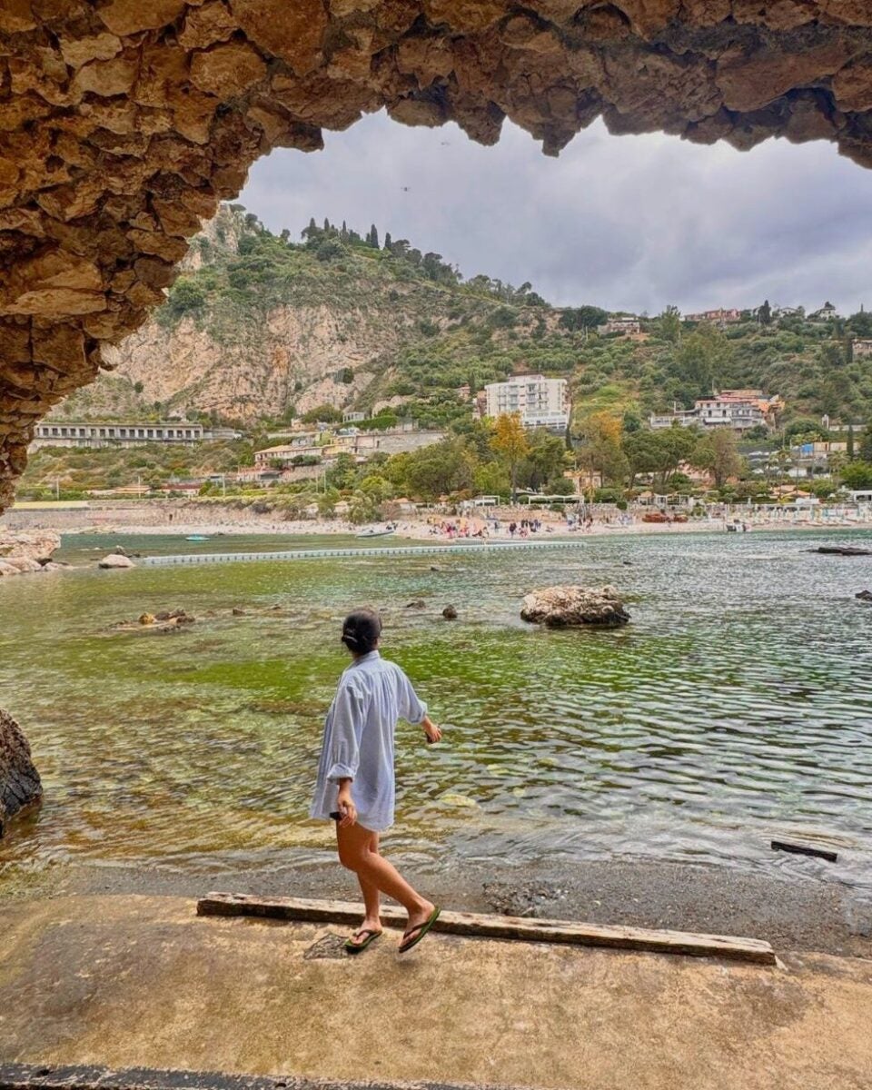 A woman standing near water under a rocky arch, with a beach and hillside in the background.