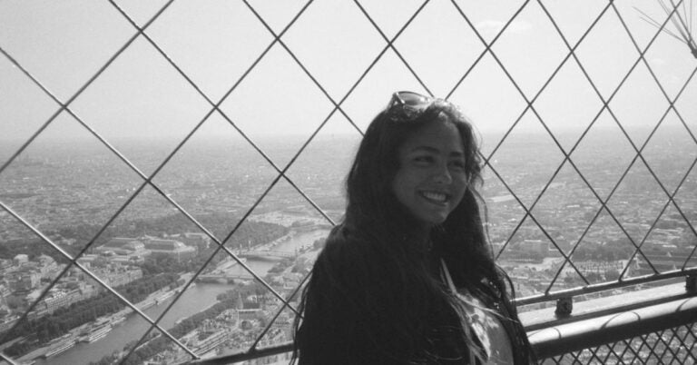 A young woman with long hair standing behind a safety net on a high observation deck overlooking a cityscape with rivers and buildings in the distance.