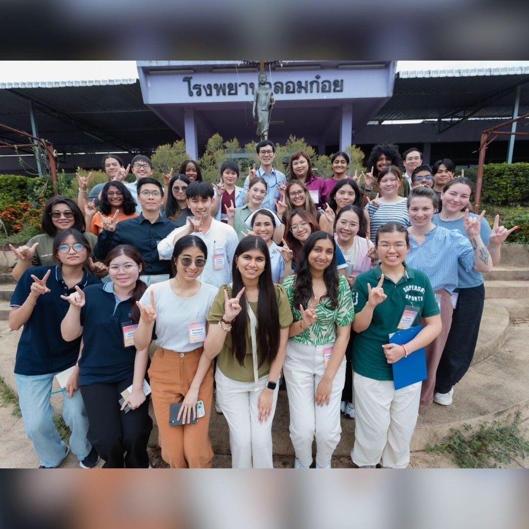 A diverse group of people, smiling and making hand gestures, stand in front of a building with Thai text.
