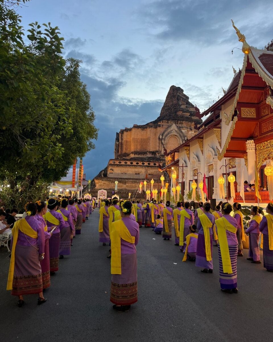 A group wearing traditional purple and yellow attire stands in formation along a street by a temple, with an ancient pagoda in the background.