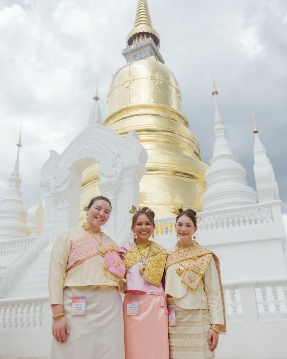 Three young women in traditional attire in front of a golden domed temple.