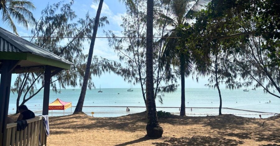 Beachfront scene with palm trees framing the view of a calm sea, small boats, and a lifeguard tent on the sand.