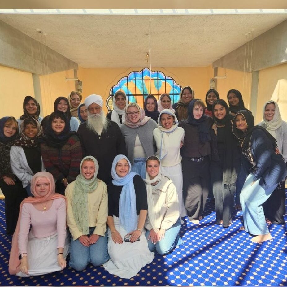 A group of people, mostly women wearing headscarves, pose together and smile for a photo inside a bright room with blue patterned carpet and arched stained glass.