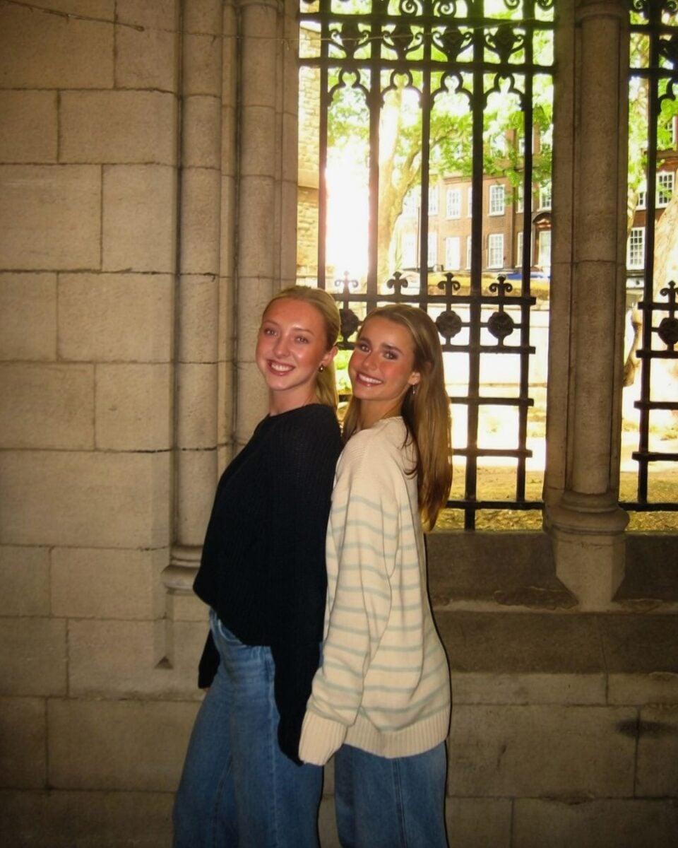 Two young women stand together in front of a large window.