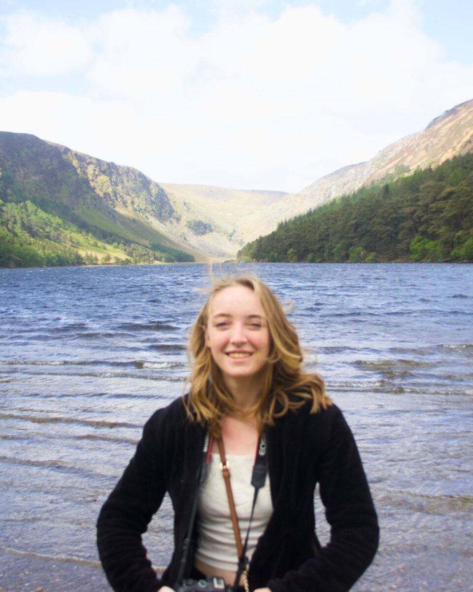 Young woman smiling in front of a serene lake surrounded by green hills and mountains under a clear sky.