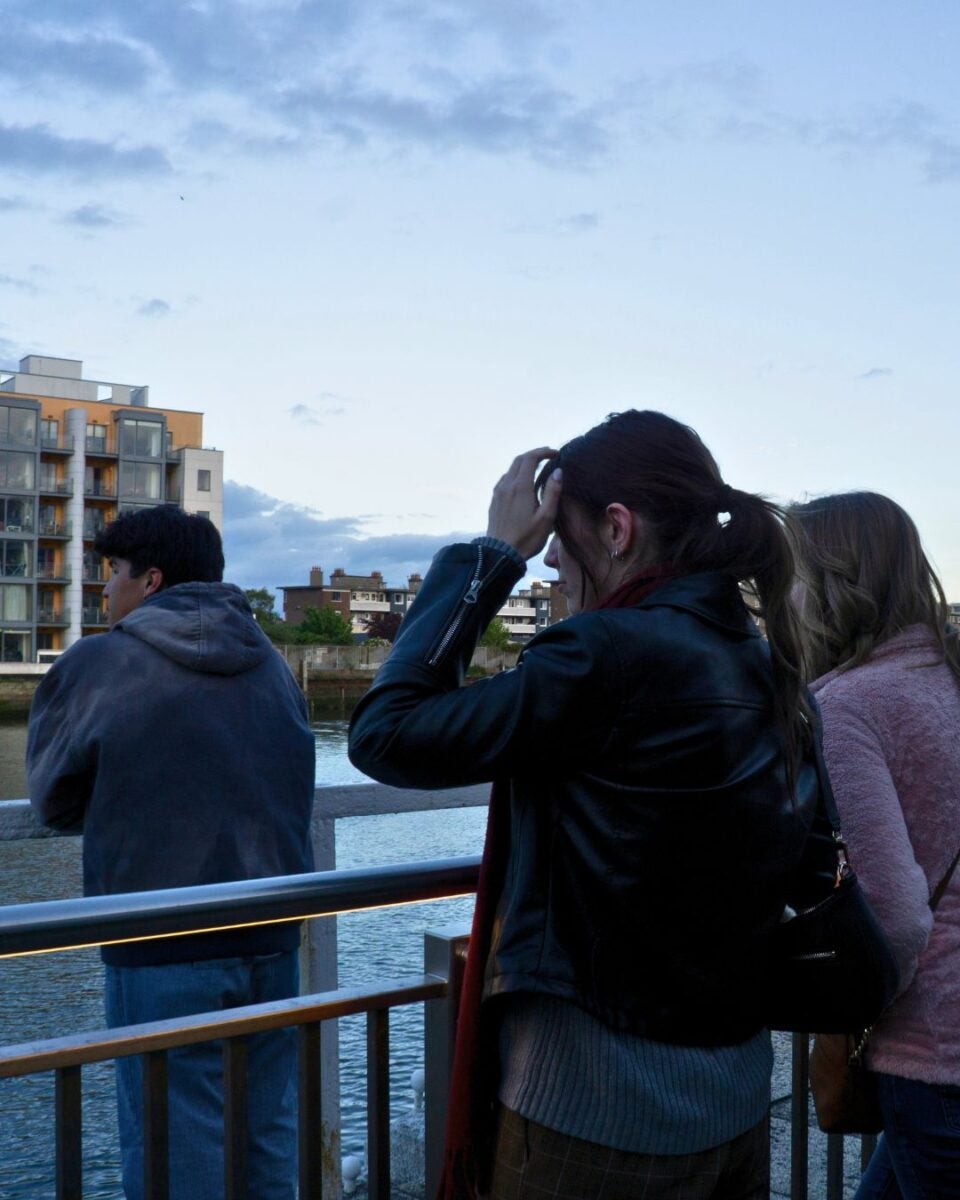 Three people stand by a waterfront railing during early evening, looking out over the water with buildings in the background.