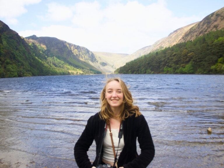 Young woman smiling in front of a serene lake surrounded by green hills and mountains under a clear sky.