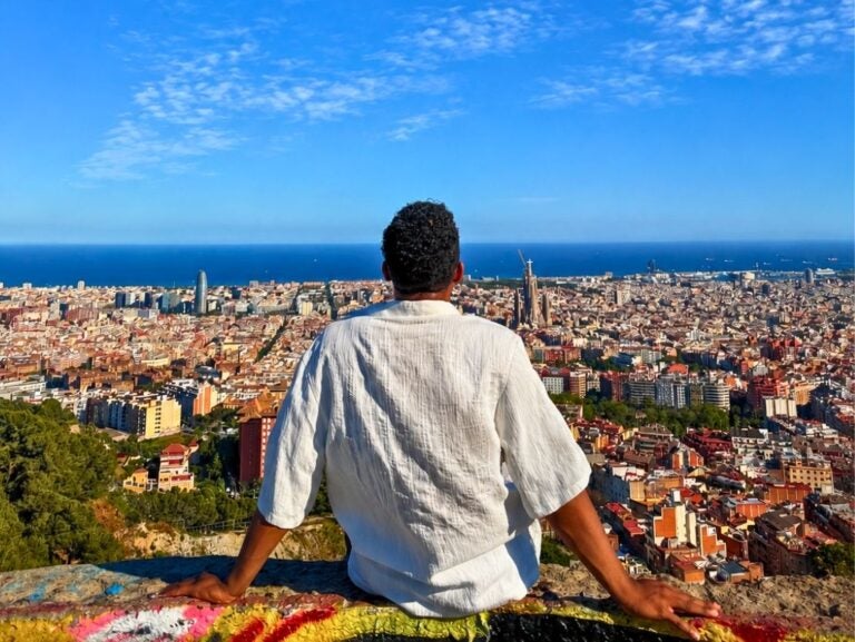 Man in white shirt sitting on a ledge overlooking a sprawling cityscape under a bright blue sky.