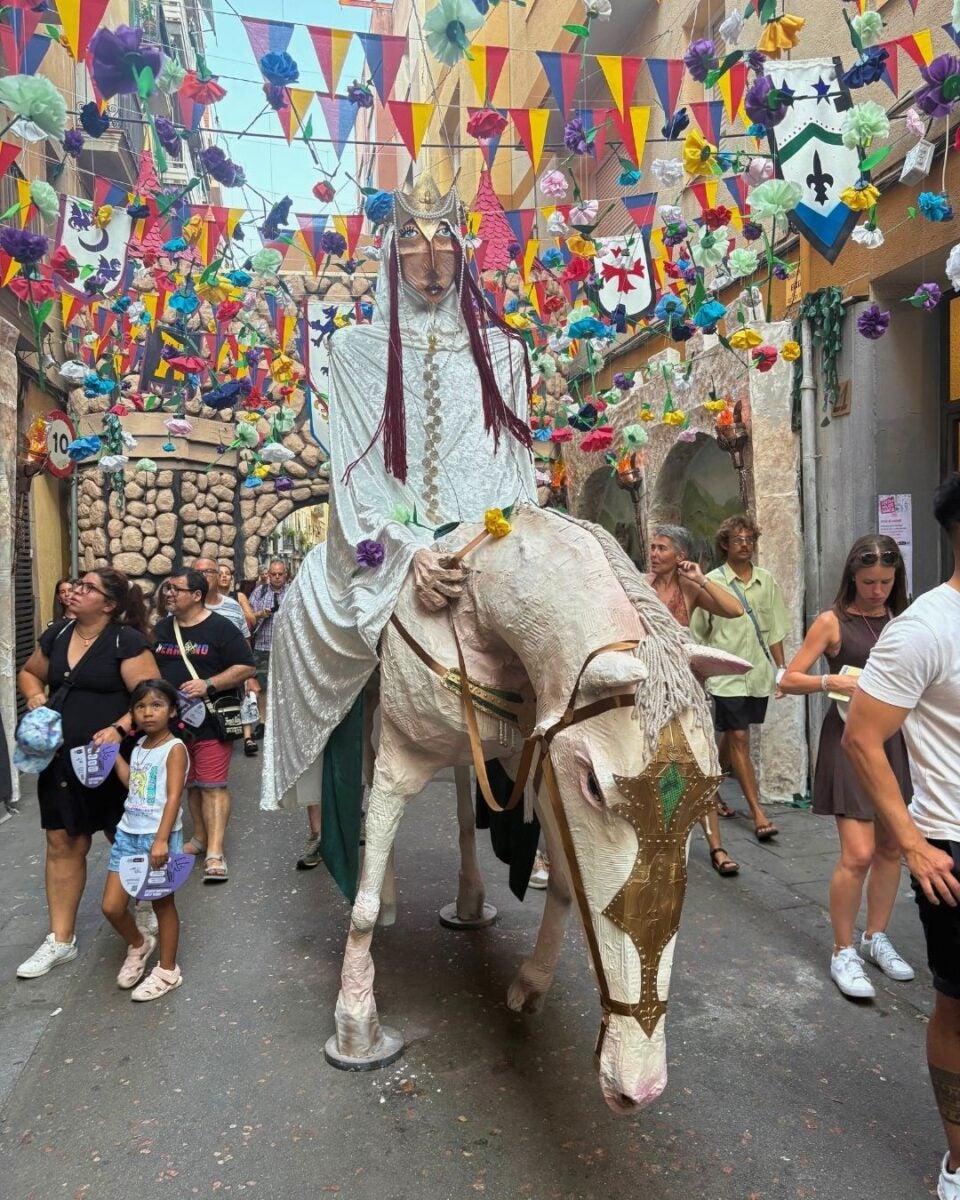 A large figure dressed in white riding a decorated horse puppet through a festive street adorned with colorful flags and flowers.