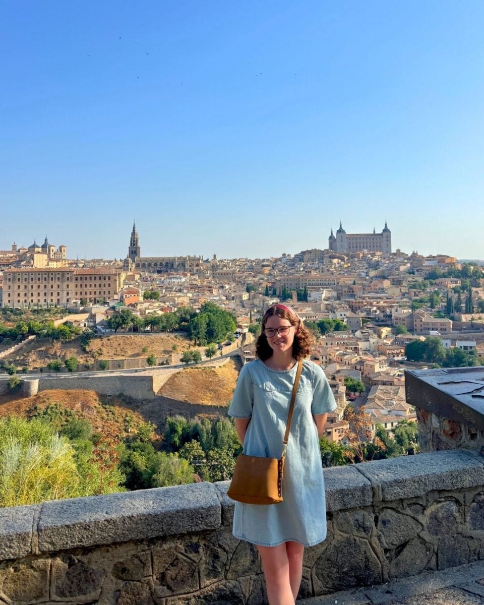 Woman in light blue dress standing on a stone terrace overlooking a historic city with a castle and church towers under a clear blue sky.