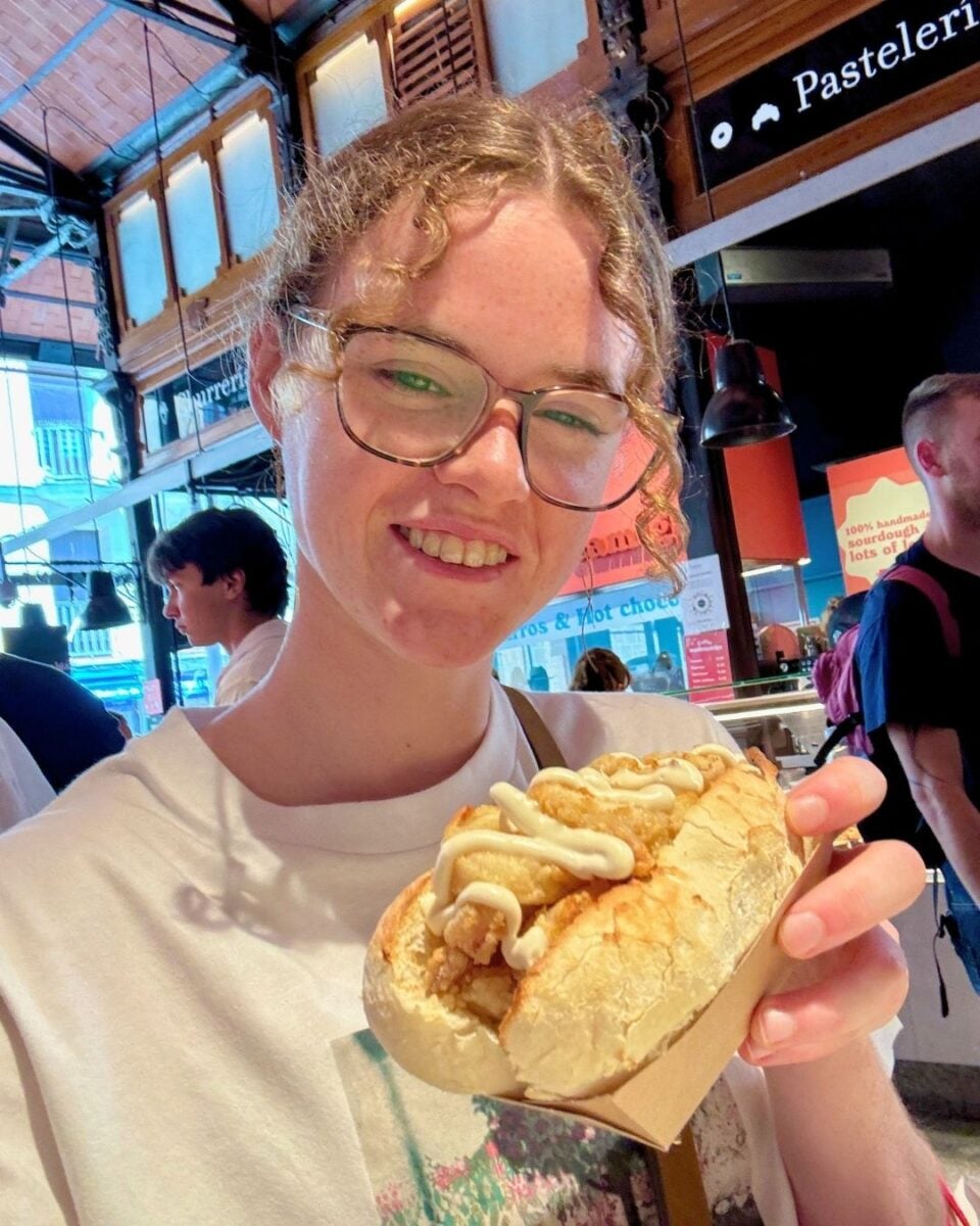 Young woman with glasses smiling and holding a sandwich topped with sauce inside a bustling indoor market.