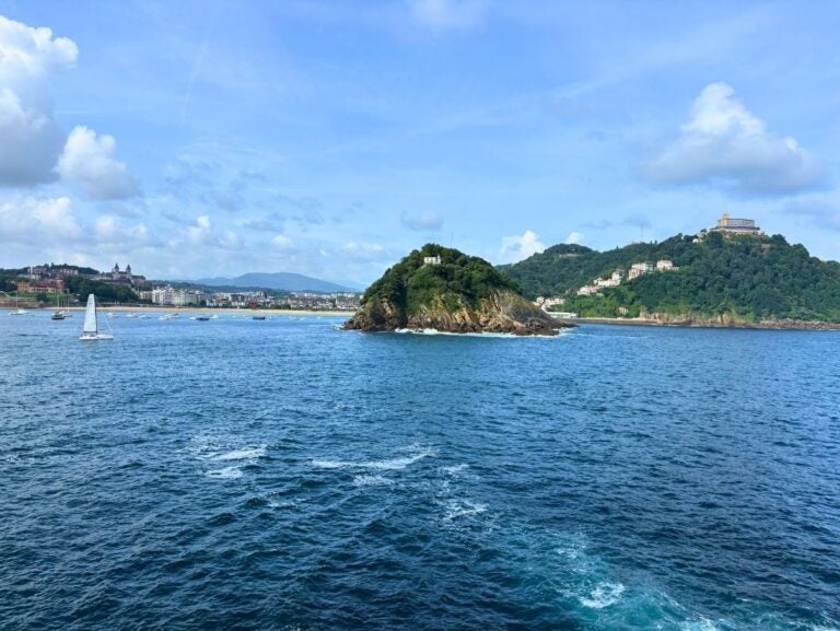 Small rocky island covered with greenery and a white building, surrounded by blue ocean waters under a partly cloudy sky.