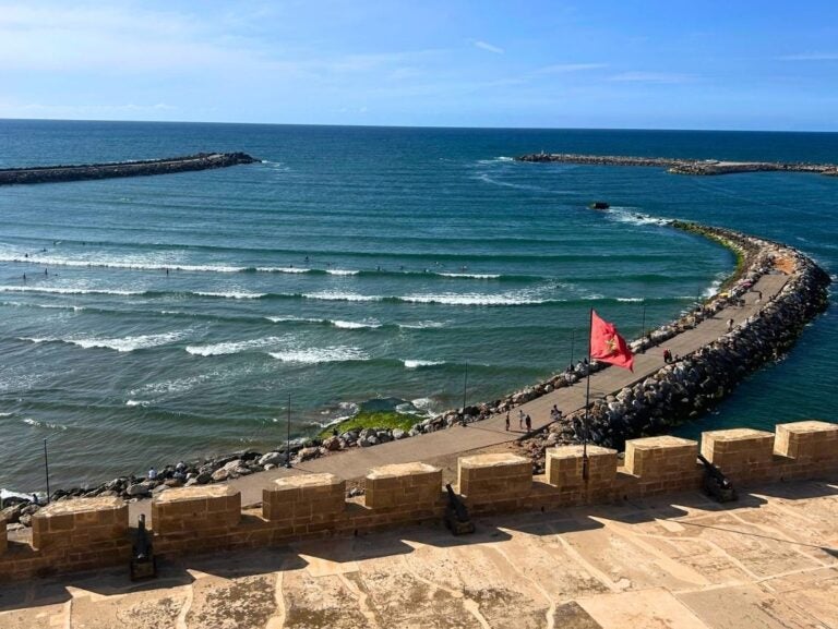 Curved stone breakwater extending into the ocean with gentle waves and a red flag flying near the shore under a clear blue sky.