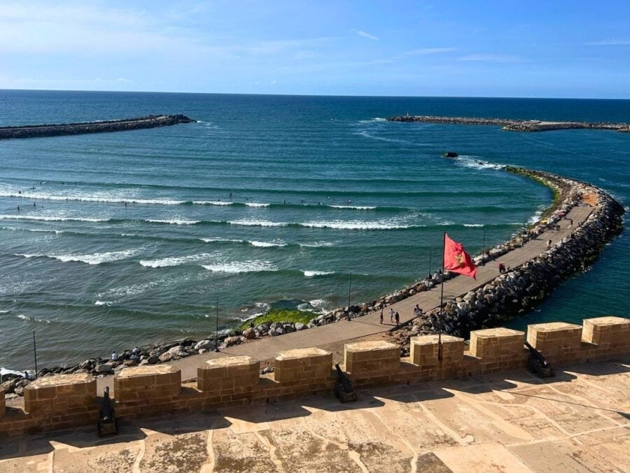 Curved stone breakwater extending into the ocean with gentle waves and a red flag flying near the shore under a clear blue sky.