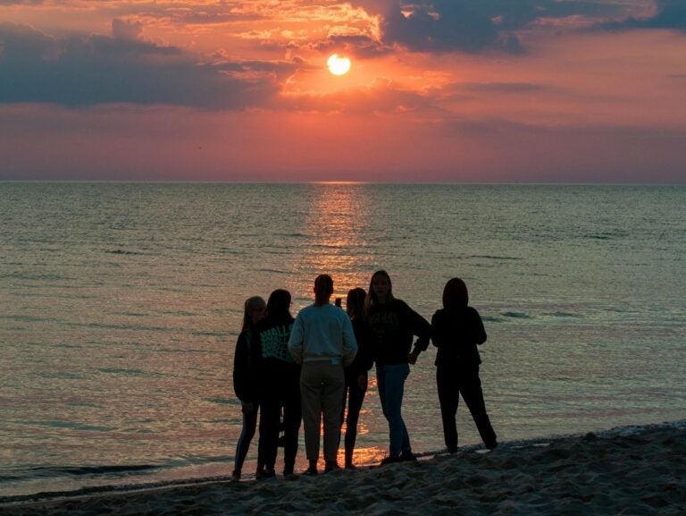 Group of six people standing on a beach watching the sun set over calm ocean waters.