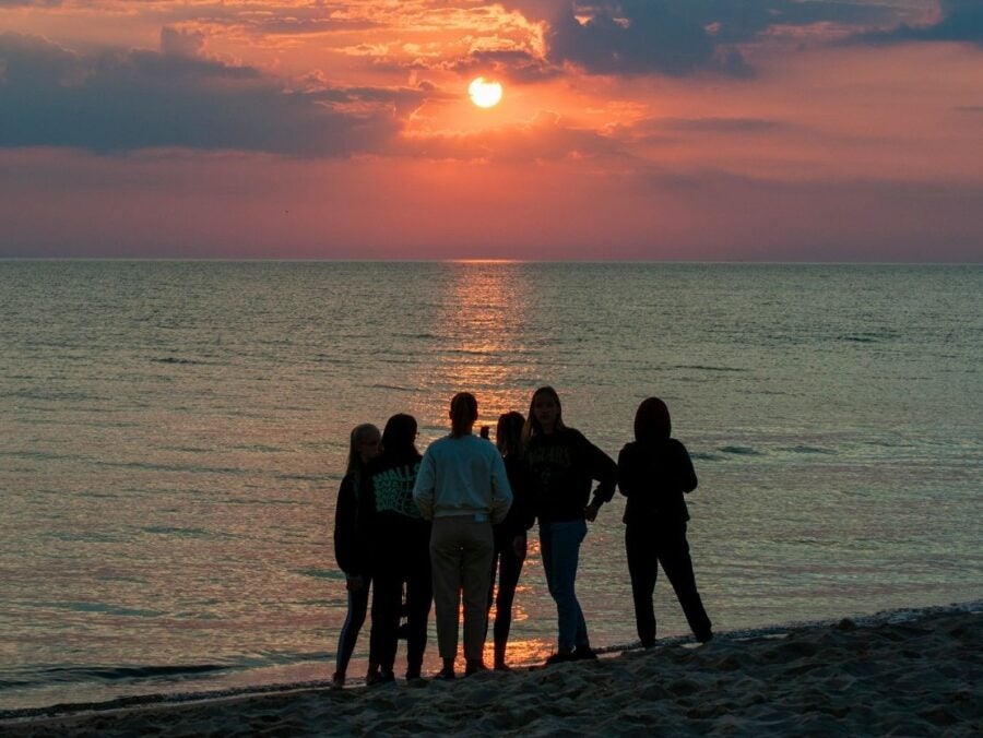 Group of six people standing on a beach watching the sun set over calm ocean waters.