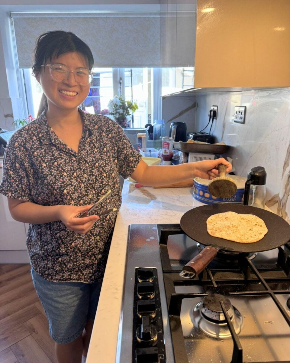 Smiling young woman cooking flatbread on a stovetop griddle in a bright kitchen with natural light.