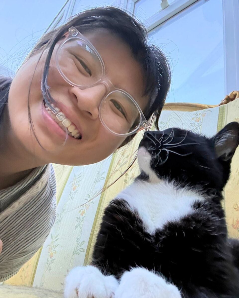 Smiling woman with glasses taking a close selfie next to a black and white cat resting on a floral patterned couch.