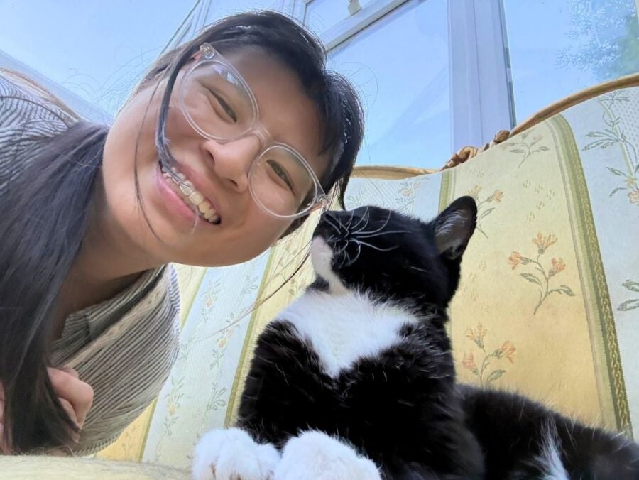 Smiling woman with glasses taking a close selfie next to a black and white cat resting on a floral patterned couch.