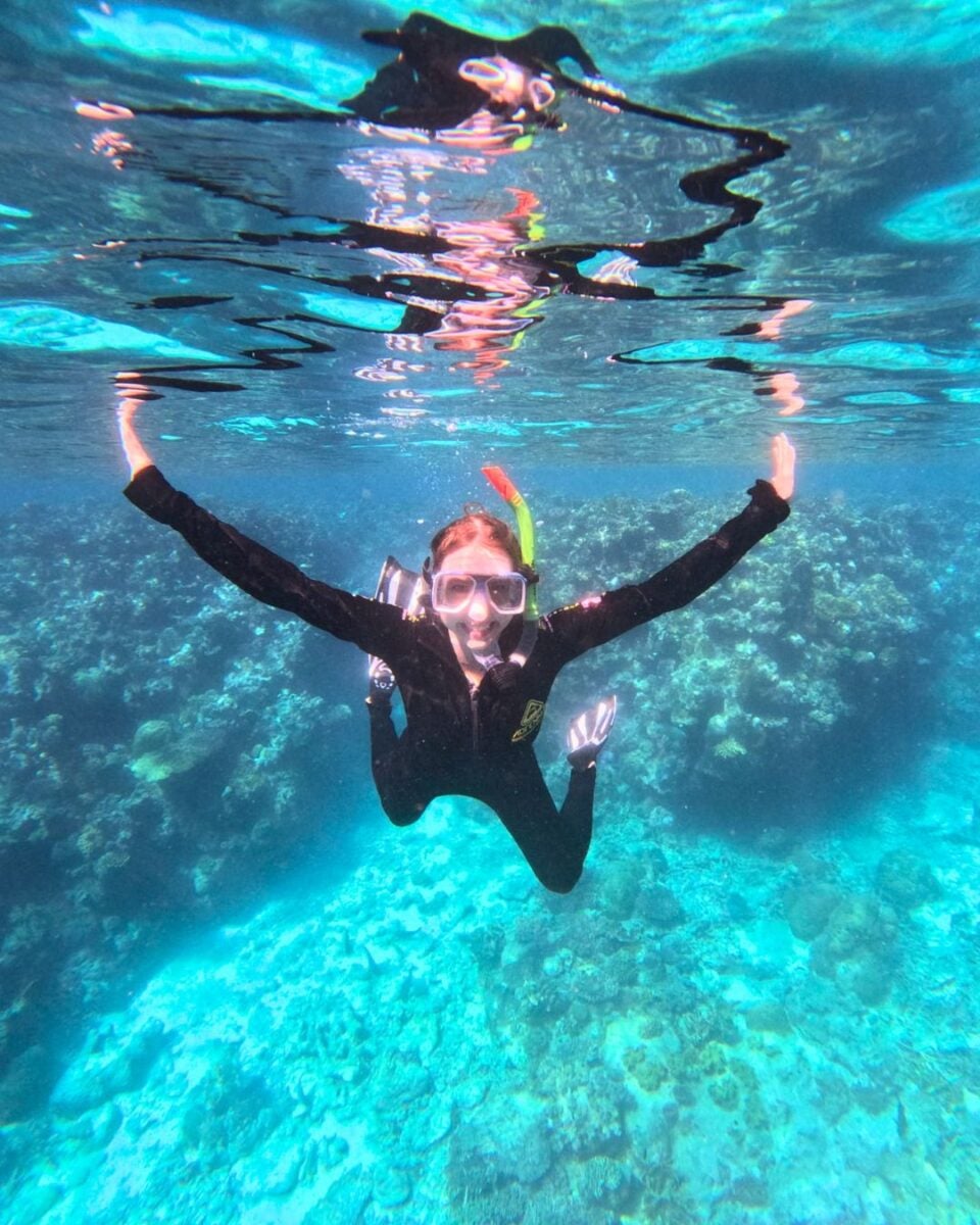 Snorkeler in black wetsuit and mask swimming underwater above a vibrant coral reef in clear blue ocean water.