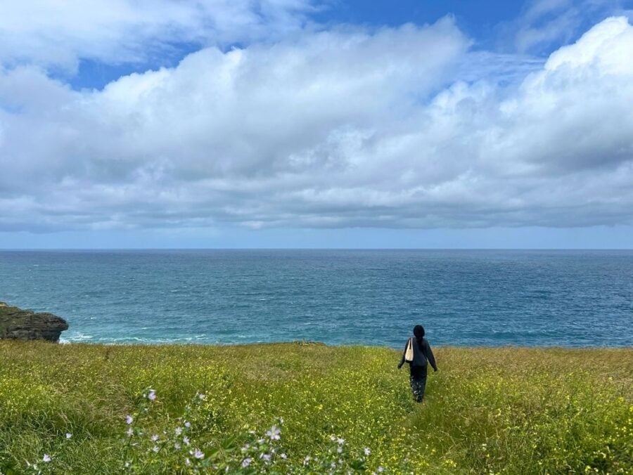 A young woman walking through a grassy field toward a vast ocean under a partly cloudy sky.