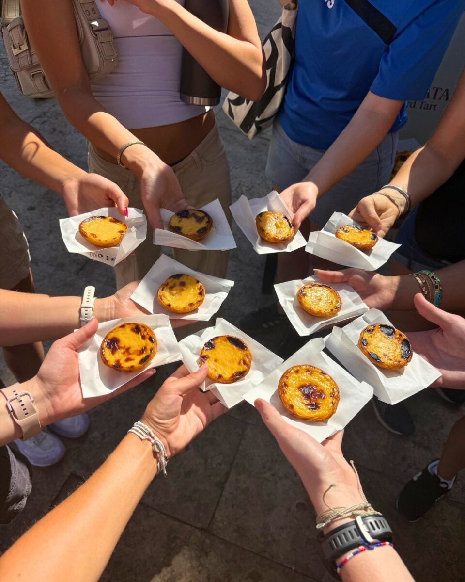 Group of people holding pastel de nata pastries on white napkins in a sunny outdoor setting.