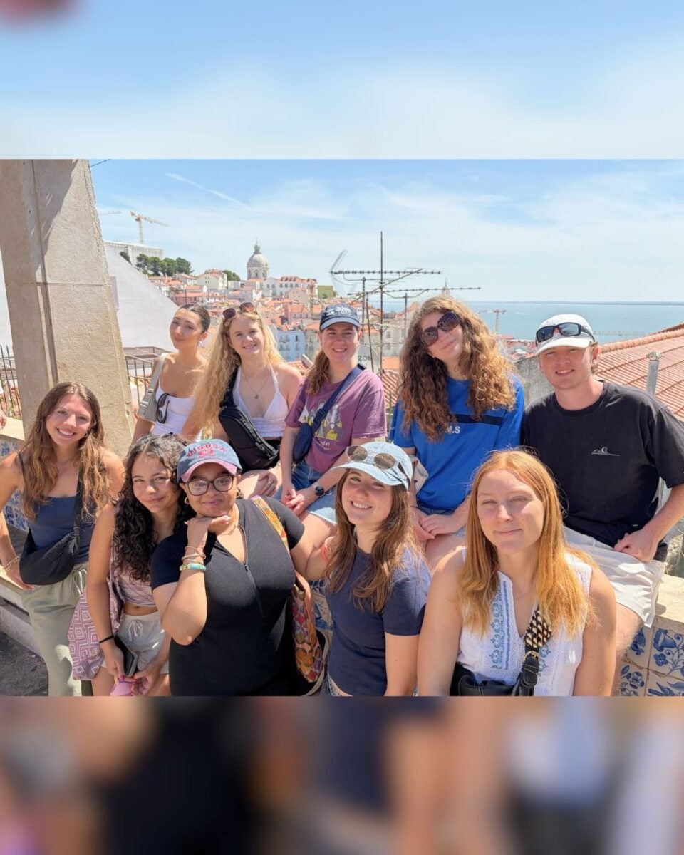 Group of nine friends posing on a sunny rooftop with a scenic city and ocean view in the background.