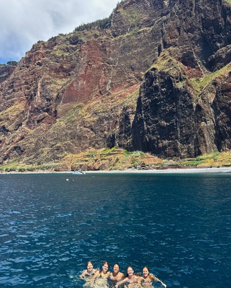 Five young women swimming in clear blue ocean water near a towering rocky cliff with a small beach at its base under a partly cloudy sky.
