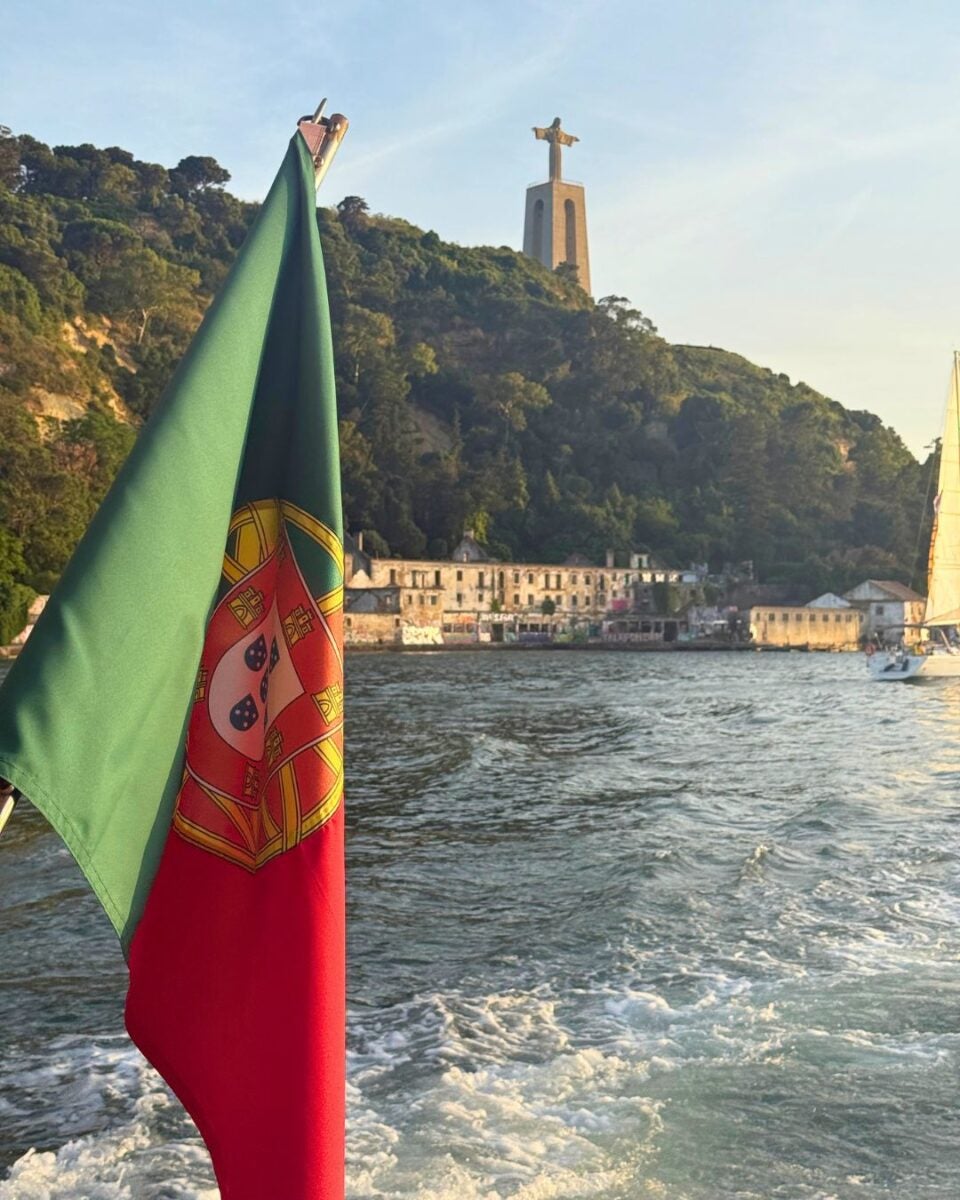 The Portugal flag waving on a boat with the Cristo Rei statue and lush hillside in the background at sunset.