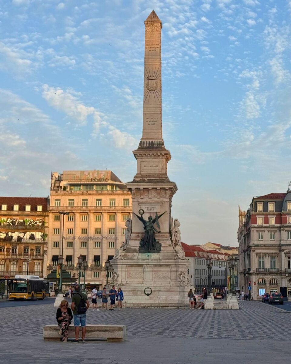 Monumento aos Restauradores. Tall stone obelisk monument with statues at its base in a bustling city square during sunset.