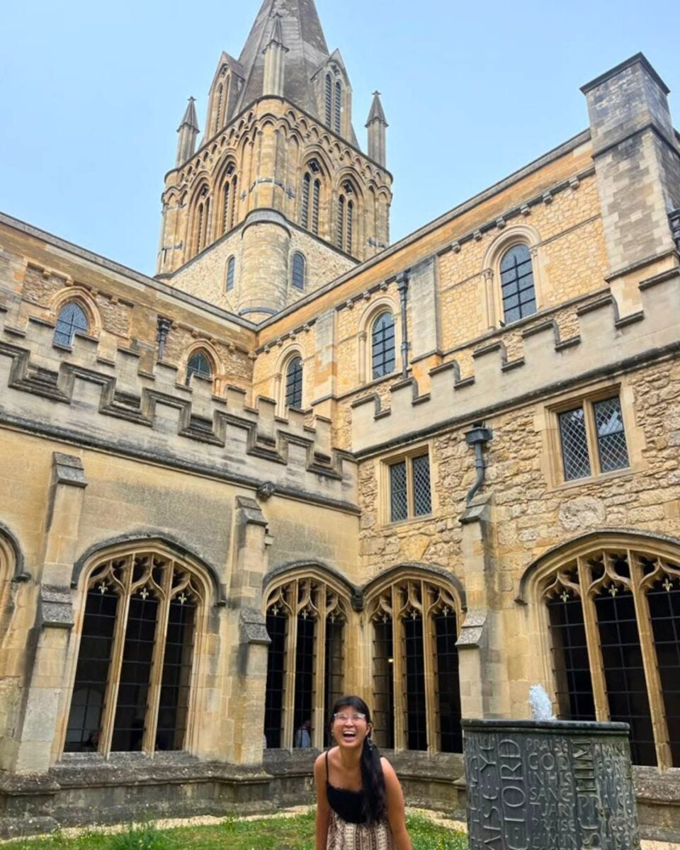 The Christ Church Cathedral building with arched windows and a tall spire under a clear sky, featuring a smiling woman in the courtyard.