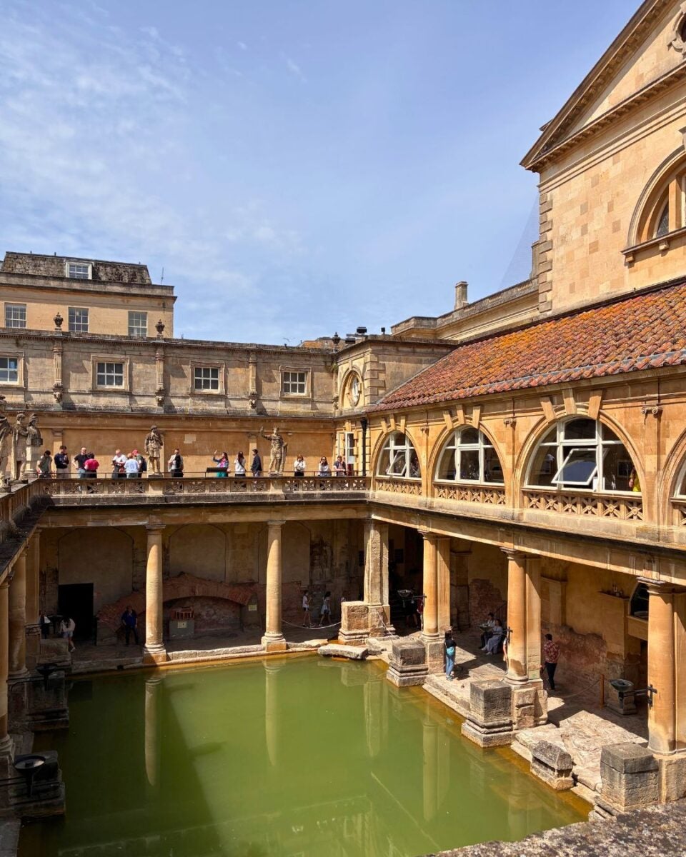 The sacred Roman Baths with green water pool surrounded by sandstone arches and columns under a clear blue sky.
